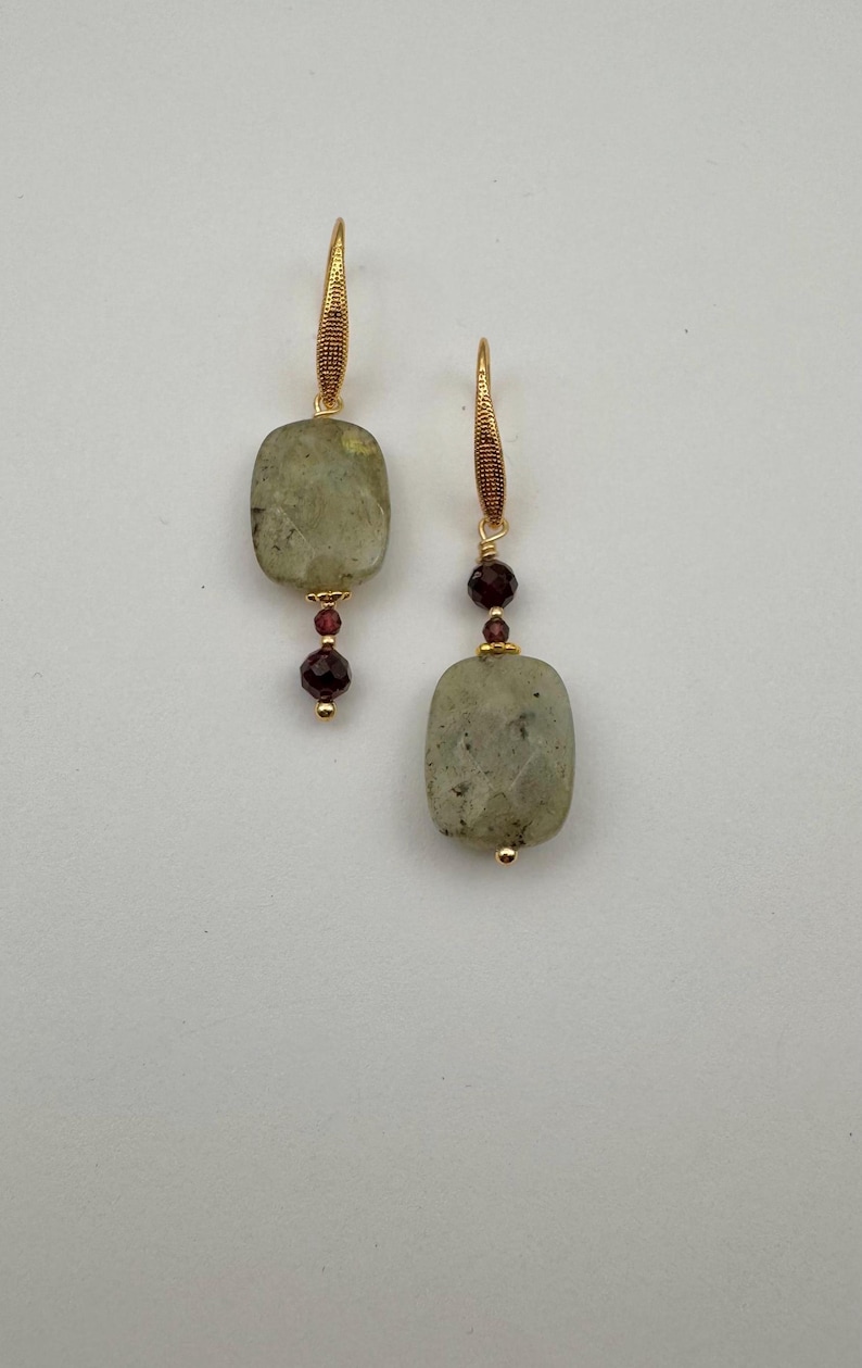 May include: A pair of gold earrings with a rectangular green stone and a small red bead. The earrings are hanging from a gold hook.