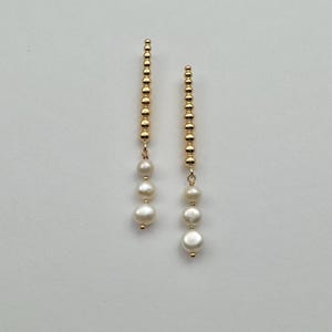 May include: A pair of gold-colored drop earrings. Each earring features a vertical strand of small, round beads, with three white, round pearls suspended below. The earrings are displayed against a plain, light background.