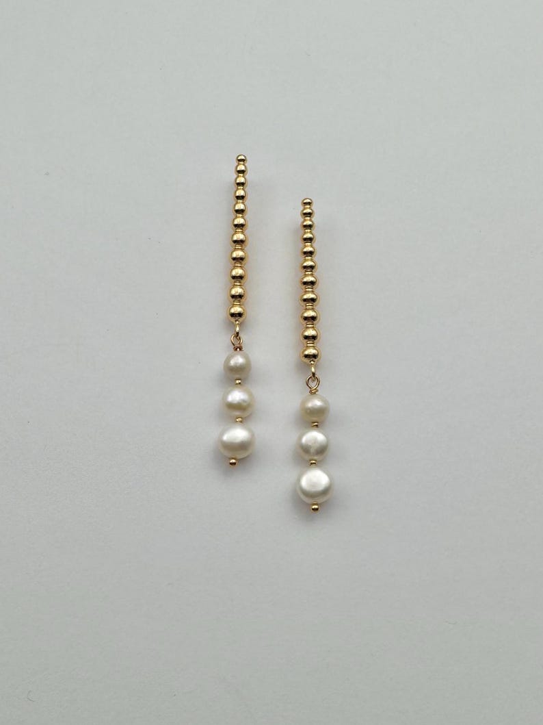 May include: A pair of gold-colored drop earrings. Each earring features a vertical strand of small, round beads, with three white, round pearls suspended below. The earrings are displayed against a plain, light background.