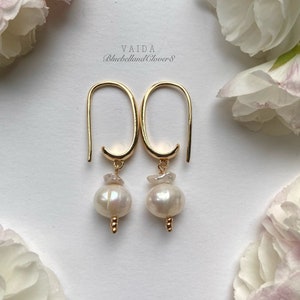 Peut inclure: Une paire de boucles d'oreilles dorées au design unique. Chaque boucle d'oreille présente un dessus incurvé, un petit groupe de pierres blanches et une grosse perle blanche ronde. Les boucles d'oreilles sont posées sur un fond blanc avec des fleurs blanches et roses. Le texte "VAIDA BluebellandClover8" est visible.