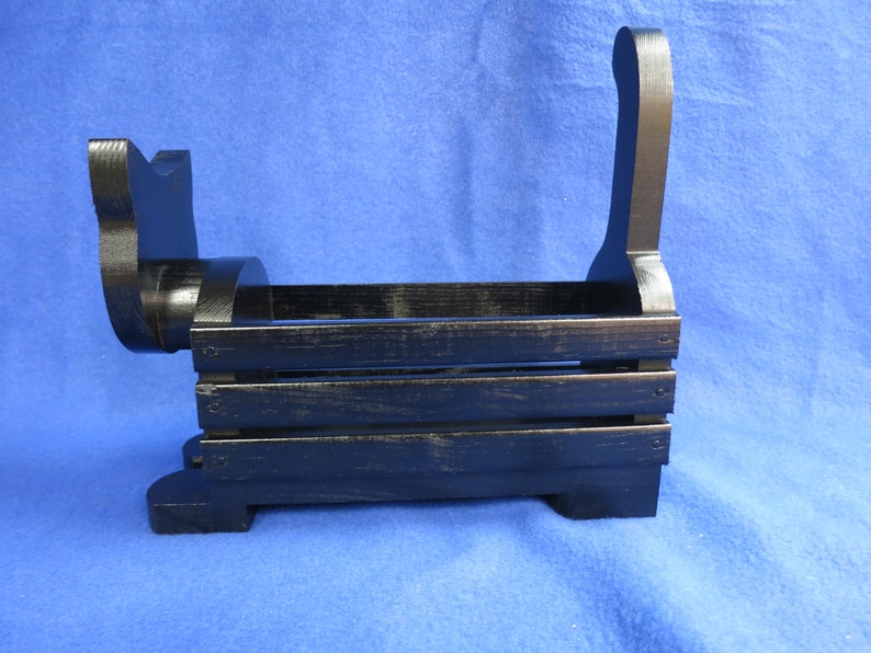 May include: Black wooden dog-shaped storage basket. The basket has a rectangular body with horizontal slats and a curved handle. The dog's head and tail are also part of the design. The basket is painted black with a distressed finish.