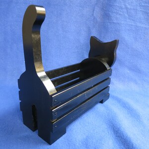 May include: A black wooden cat-shaped basket with a handle shaped like a cat's tail. The basket has a rectangular body with horizontal slats and a cat head silhouette at the back. The cat basket is painted black.