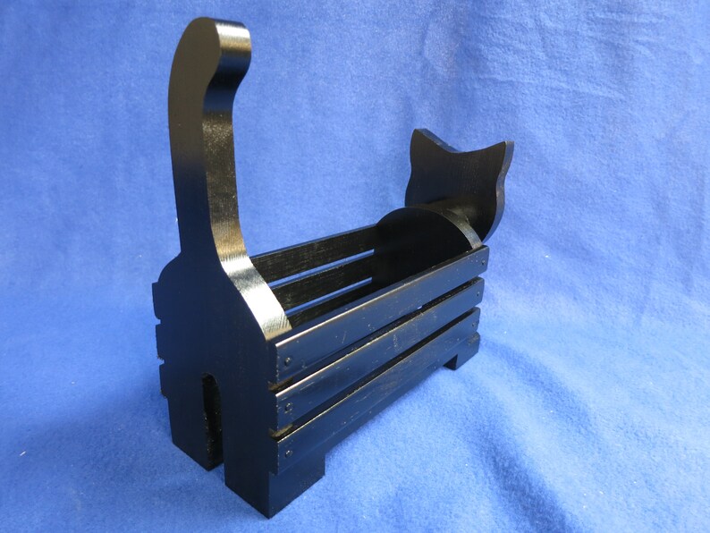 May include: A black wooden cat-shaped basket with a handle shaped like a cat's tail. The basket has a rectangular body with horizontal slats and a cat head silhouette at the back. The cat basket is painted black.
