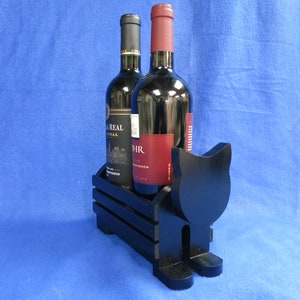 May include: A black wooden cat-shaped wine bottle holder with two bottles of red wine. The holder has a crate-like design to hold the bottles. The bottles have black and red labels. The background is a solid blue.