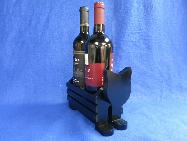May include: A black wooden cat-shaped wine bottle holder with two bottles of red wine. The holder has a crate-like design to hold the bottles. The bottles have black and red labels. The background is a solid blue.