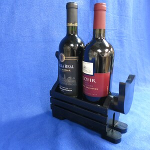 May include: A black wooden wine bottle holder with two wine bottles. One bottle has a black cap and a gold label, the other has a red cap and a red label. The holder is shaped like a dog.