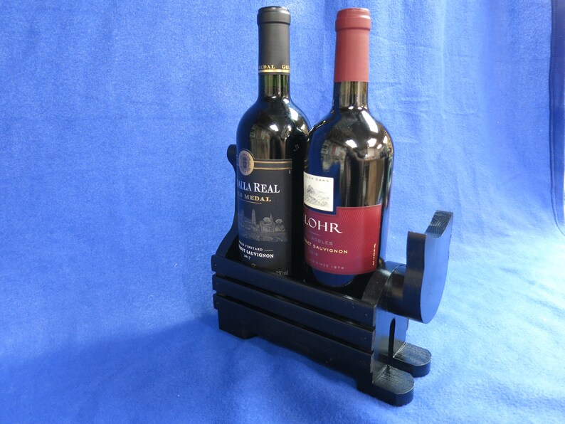 May include: A black wooden wine bottle holder with two wine bottles. One bottle has a black cap and a gold label, the other has a red cap and a red label. The holder is shaped like a dog.