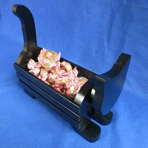 May include: A black wooden sleigh-shaped candy dish filled with wrapped candies. The dish has a curved handle and runners, and the candies are in red and white wrappers. The dish is on a blue surface.