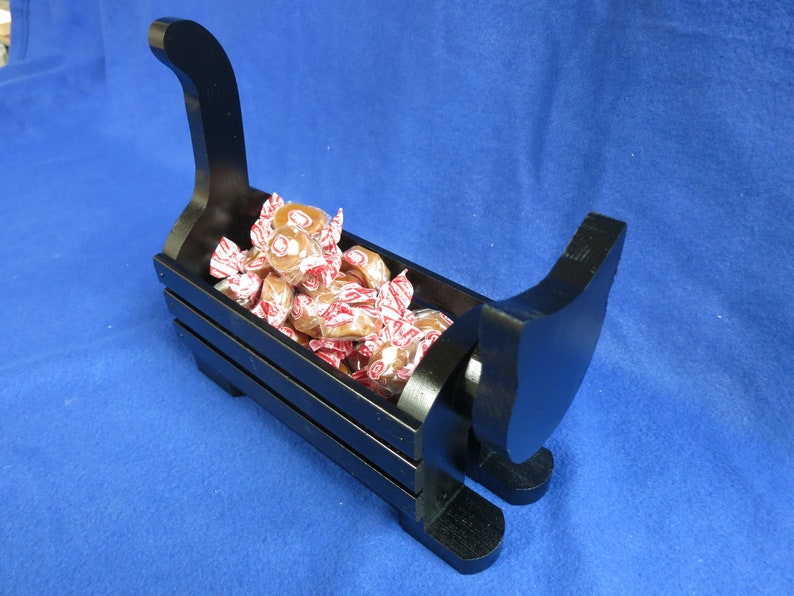 May include: A black wooden sleigh-shaped candy dish filled with wrapped candies. The dish has a curved handle and runners, and the candies are in red and white wrappers. The dish is on a blue surface.