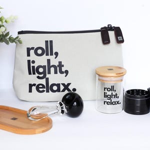 Pode incluir: Um conjunto de acessórios para relaxamento, incluindo uma bolsa de lona com o texto "roll, light, relax", um frasco de vidro, um moedor preto, uma bandeja de madeira e um cachimbo de vidro. A bolsa e o frasco também apresentam o texto "roll, light, relax".