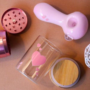 May include: A set of pink and silver smoking accessories, including a grinder, a pipe, a glass jar with a wooden lid, and a small white decorative ball.