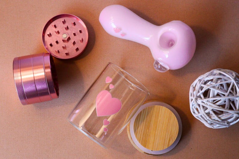 May include: A set of pink and silver smoking accessories, including a grinder, a pipe, a glass jar with a wooden lid, and a small white decorative ball.