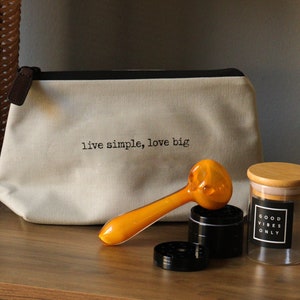 May include: A white canvas bag with the text "live simple, love big" printed on it.  A yellow glass pipe, a black grinder, and a clear glass jar with a wooden lid and the text "good vibes only" printed on a black label.