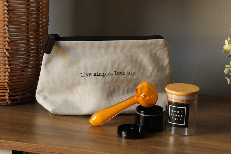 May include: A white canvas bag with the text "live simple, love big" printed on it.  A yellow glass pipe, a black grinder, and a clear glass jar with a wooden lid and the text "good vibes only" printed on a black label.
