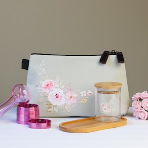 May include: A collection of accessories, including a pink glass pipe, a grinder, and a cosmetic bag. The bag and glass jar are decorated with floral designs. A small bouquet of pink roses and a wooden tray are also present.