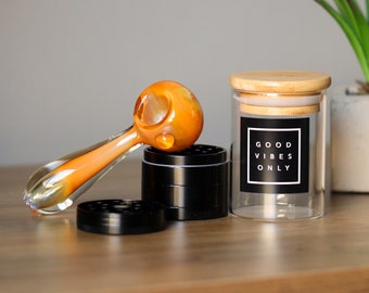 May include: A glass pipe with an orange and yellow swirl design, a black metal grinder, and a glass jar with a bamboo lid and a black label that says "Good Vibes Only".