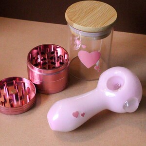 May include: A pink glass pipe with a heart design, a pink metal grinder with three parts, and a glass jar with a wooden lid and a heart design.