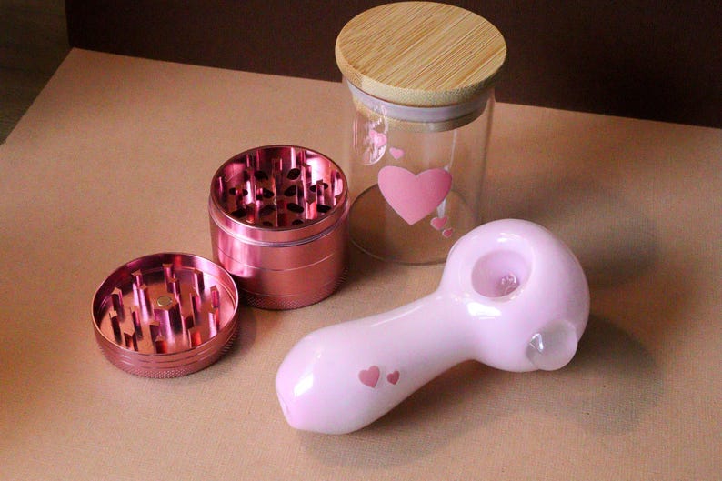 May include: A pink glass pipe with a heart design, a pink metal grinder with three parts, and a glass jar with a wooden lid and a heart design.