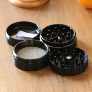 May include: Four black metal grinder pieces with a screen and a small white paper inside one of the pieces.