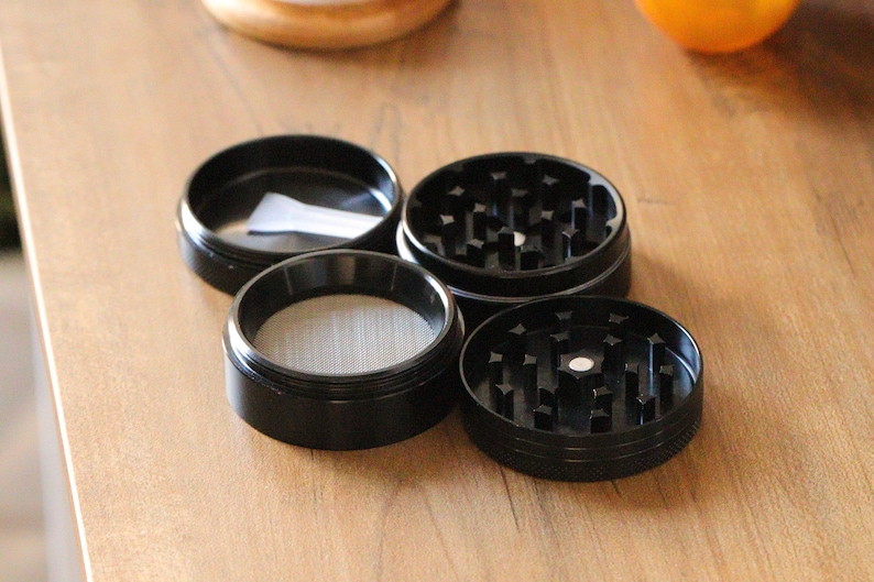 May include: Four black metal grinder pieces with a screen and a small white paper inside one of the pieces.