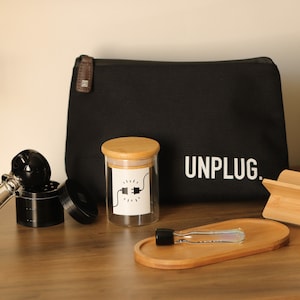 Op de afbeelding: Een zwarte canvas tas met het woord "UNPLUG." in wit gedrukt. De tas ligt naast een glazen pot met een bamboe deksel, een glazen pijp, een grinder, een houten dienblad en een houten standaard.