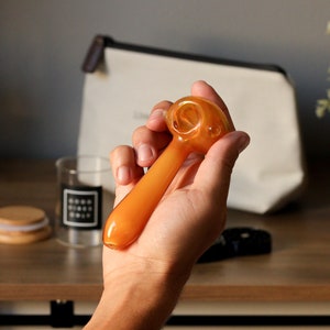 May include: A hand holding a small, amber-colored glass pipe with a round bowl. The pipe is about 5 inches long.
