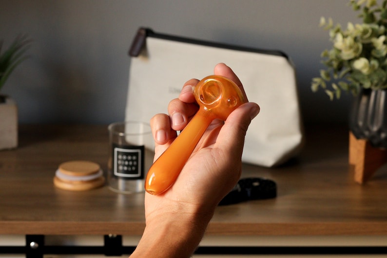 May include: A hand holding a small, amber-colored glass pipe with a round bowl. The pipe is about 5 inches long.