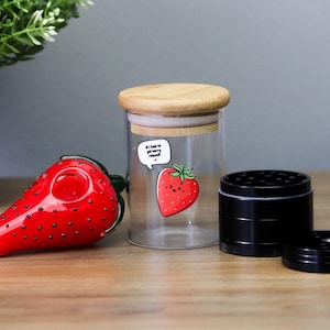 May include: A glass strawberry-shaped pipe, a glass jar with a wooden lid and a black metal grinder. The jar has a sticker with a cartoon strawberry and the text "It's time to get berry mellow".