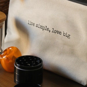 May include: A white canvas pouch with the text "live simple, love big" printed on it. The pouch is next to a glass jar with a wooden lid and the text "good vibes only" printed on a black label. There is also a glass pipe and a metal grinder.