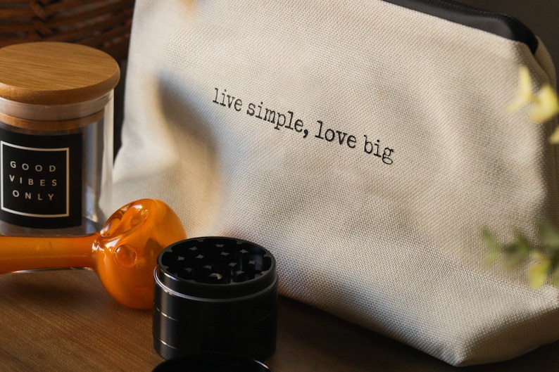 May include: A white canvas pouch with the text "live simple, love big" printed on it. The pouch is next to a glass jar with a wooden lid and the text "good vibes only" printed on a black label. There is also a glass pipe and a metal grinder.