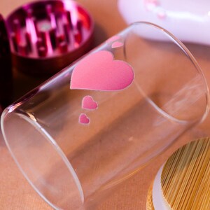 May include: A clear glass jar with a pink heart design and a bamboo lid. The jar is sitting on a brown surface.