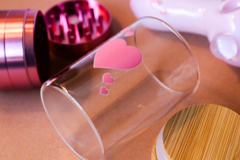May include: A clear glass jar with a pink heart design and a bamboo lid. The jar is sitting on a brown surface.
