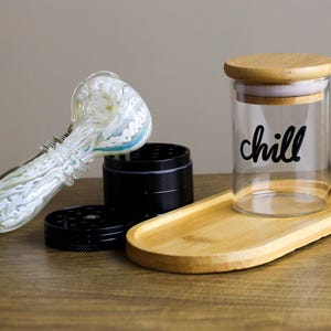 Op de afbeelding: Een glazen pijp met een wit swirl-ontwerp, een zwarte grinder en een glazen pot met een houten deksel en het woord "chill" erop. De pot staat op een houten dienblad.