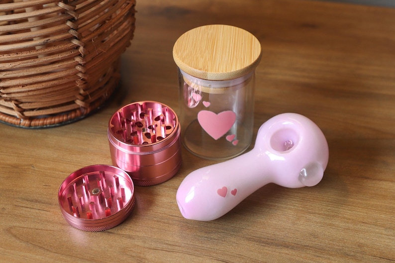 May include: A pink glass pipe with a heart design, a pink metal grinder with a three-piece design, and a glass jar with a wooden lid and a heart design.