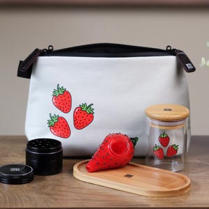 Puede incluir: Un conjunto de accesorios con temática de fresas. Incluye una bolsa blanca con gráficos de fresas, un tarro de cristal con tapa de madera, una pipa roja con forma de fresa, un molinillo negro y una bandeja de madera.