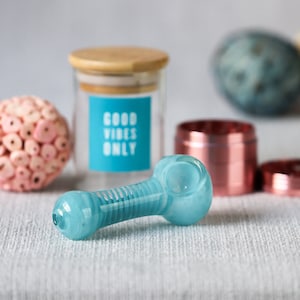 Op de afbeelding: Een lichtblauwe glazen pijp met een geribbeld ontwerp. De pijp ligt op een grijs, getextureerd oppervlak. Op de achtergrond staat een glazen pot met een houten deksel. De pot heeft een blauw etiket met de tekst "Good Vibes Only".