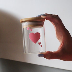May include: A clear glass jar with a wooden lid and pink heart stickers. The jar is held in a hand.