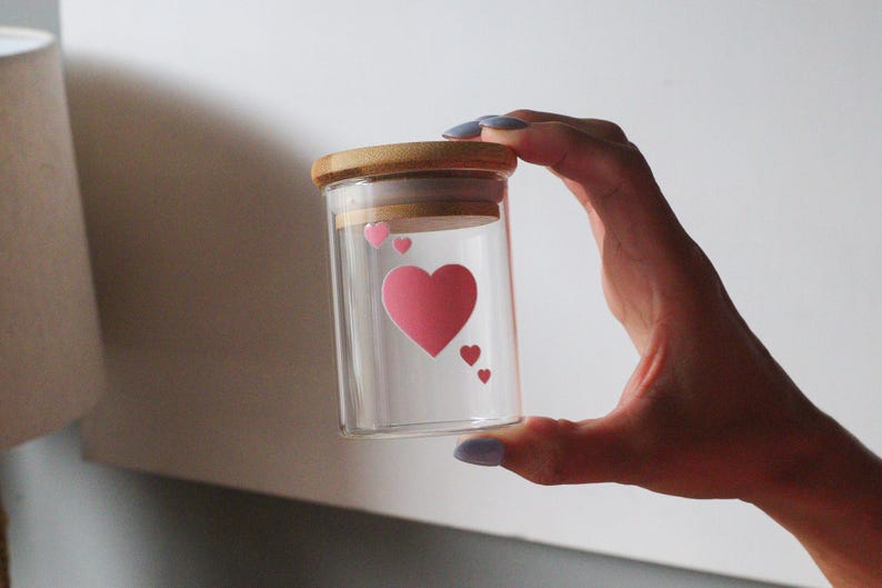May include: A clear glass jar with a wooden lid and pink heart stickers. The jar is held in a hand.
