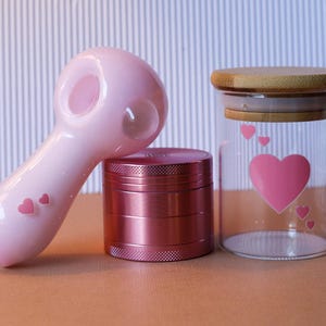 May include: A pink glass pipe with a heart design, a pink metal grinder, and a glass jar with a wooden lid and a heart design.