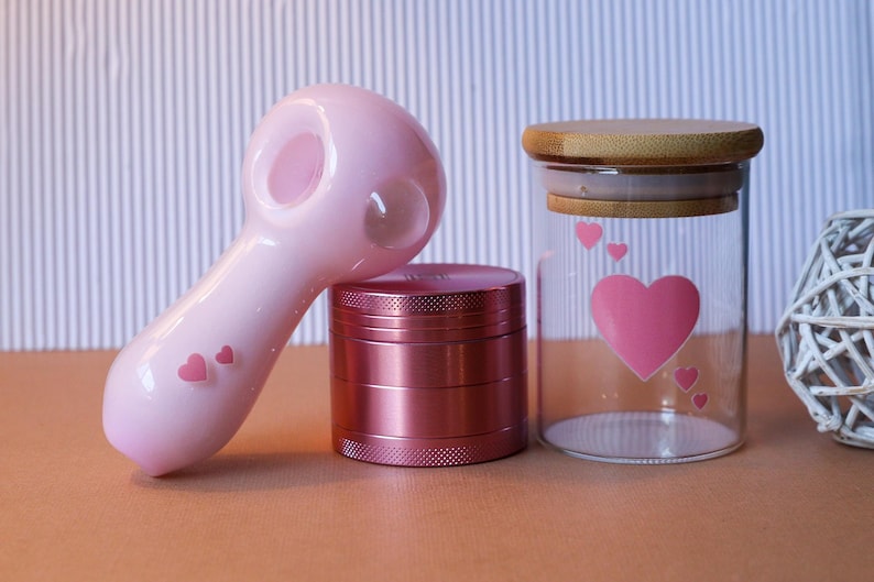 May include: A pink glass pipe with a heart design, a pink metal grinder, and a glass jar with a wooden lid and a heart design.