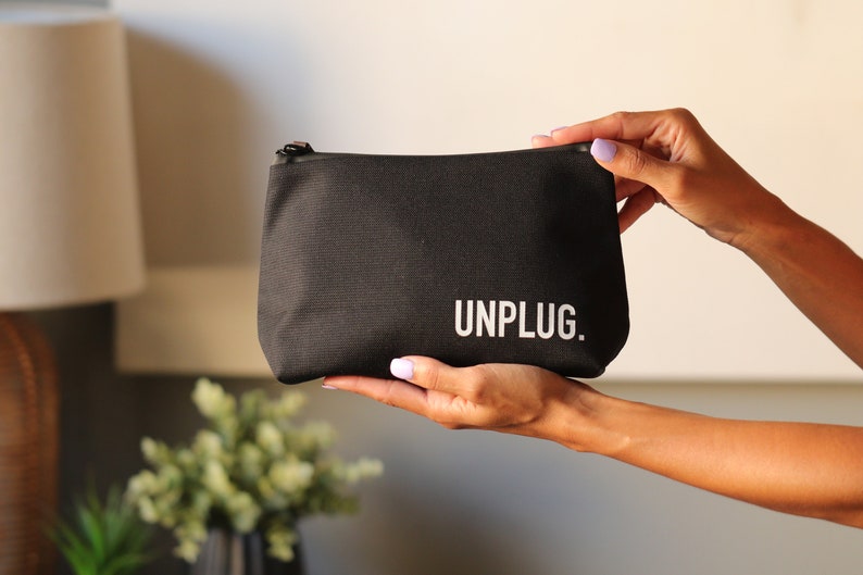 Może przedstawiać: Czarna kosmetyczka z zamkiem błyskawicznym z nadrukiem "UNPLUG." w kolorze białym.