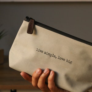 May include: A beige canvas zippered pouch with a brown leather zipper pull. The pouch has black trim and the text "live simple, love big" printed on the front.