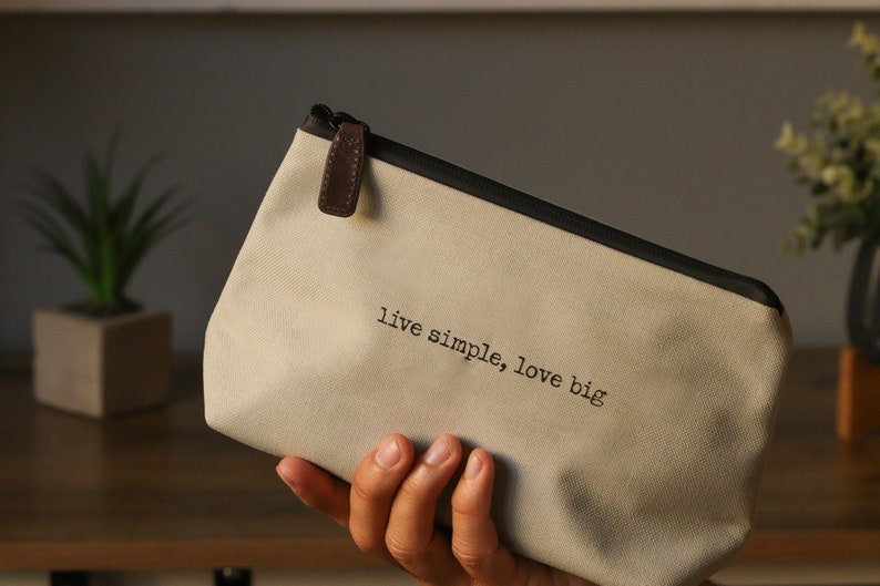 May include: A beige canvas zippered pouch with a brown leather zipper pull. The pouch has black trim and the text "live simple, love big" printed on the front.