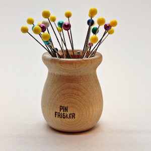 May include: A wooden pin holder with the text "PIN FRISKER" on the front, filled with colorful pins. The pins have yellow, pink, blue, and green spherical heads. The holder is made of light-colored wood.
