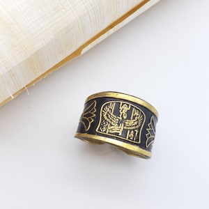 May include: A black and gold ring with Egyptian hieroglyphic designs. The ring is on a white surface next to a rolled-up piece of papyrus paper. The ring has a gold border and black center with gold hieroglyphs.