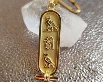 May include: Gold pendant necklace featuring an Egyptian hieroglyphic cartouche with a winged scarab beetle and an ankh symbol.