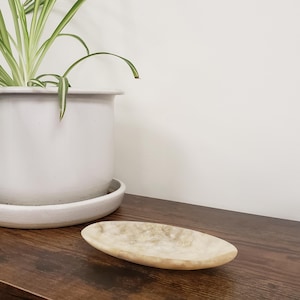 May include: A decorative, oval-shaped, beige marble tray sits on a dark brown wooden surface. A white ceramic pot with a green plant is in the background. The tray is approximately 10 inches long, perfect for displaying small items.