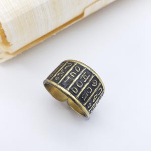 May include: A bronze-colored ring with black hieroglyphic symbols. The ring is open at the bottom and sits on a white surface, with a rolled papyrus scroll in the background.