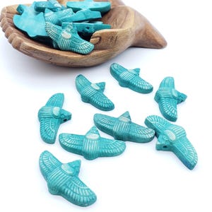 May include: A collection of turquoise-colored, winged scarab beetle beads with detailed etched wing patterns. Several beads are scattered on a white surface, while others are contained within a carved wooden bowl shaped like two cupped hands.