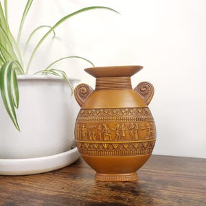 May include: A brown decorative vase with an ancient Egyptian design. The vase has a wide base, a rounded body, and two scrolled handles. The body features detailed relief carvings of figures and hieroglyphs. A white potted plant is in the background.